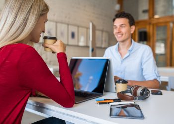 young people sitting at table face to face, working at laptop in co-working office, man smiling, startup team, freelancers, modern open space, workplace, students learning, drinking coffee