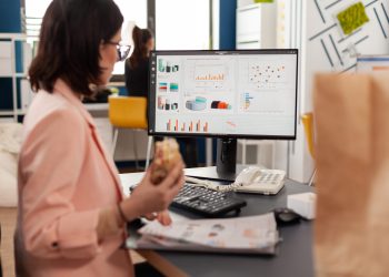 Businesswoman eating tasty sandwich having meal break working in business company office during takeout lunchbreak. Fast food order paper bag delivered at workplace. Woman typing strategy on computer.